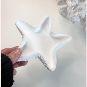 May include: A white, starfish-shaped stoneware dish with a textured rim. The dish is held by a hand. The words "ECO-FRIENDLY STONEWARE" and "SUSTAINABLE HOME DECOR" are printed vertically on either side of the image.