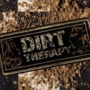May include: A rectangular black embroidered patch with a gold border and the text DIRT THERAPY in gold lettering. The patch features a textured, distressed appearance and is positioned on a black and white checkered surface covered in brown mud.