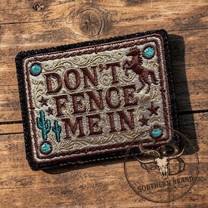 May include: A rectangular embroidered patch with a black border featuring the text DON'T FENCE ME IN in dark brown letters. The design includes a brown horse, two teal cacti, and small teal circles on a cream-colored background with swirling patterns.