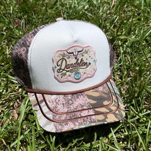 May include: A trucker hat with a white front panel and a camouflage print on the mesh back and brim. The front features an embroidered patch with the word Dandelion, a bull skull, floral accents, and a turquoise-colored stone detail.