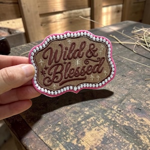 May include: An embroidered patch with a tan center featuring the dark brown text Wild & Blessed, accented by small embroidered stars. The patch has a scalloped border outlined in white and a bright pink outer edge, shaped like a western buckle.