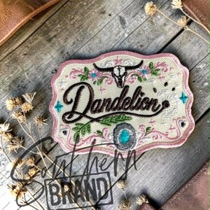 May include: A rectangular embroidered patch with scalloped edges featuring the word Dandelion in dark brown script. The design includes a black steer skull, pink floral accents, small teal stars, and a central oval turquoise stone setting.