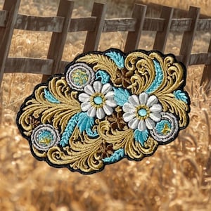 May include: An oval-shaped embroidered patch featuring two white daisies with light blue centers, surrounded by intricate gold and brown filigree scrollwork, teal leaf accents, and circular silver-toned borders, all outlined in black thread.