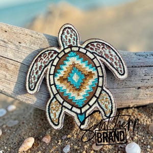 May include: An embroidered sea turtle patch featuring a geometric pattern on its shell in shades of turquoise, brown, and cream. The turtle has light blue and brown patterned flippers with a dark brown border. The patch is shaped like a sea turtle.
