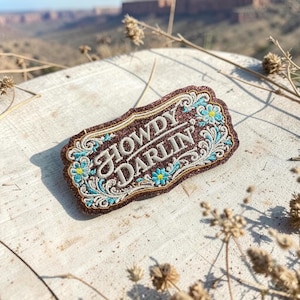 May include: A brown, glittery patch with the words "HOWDY DARLIN" in white script. The patch has a decorative border and features blue and yellow floral accents. The patch is rectangular with a curved top and bottom.