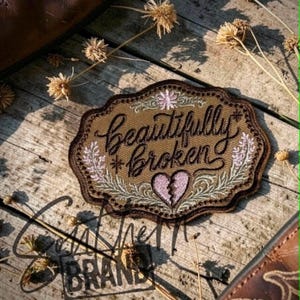 May include: An embroidered patch with a brown background and a dark brown scalloped border. The text "beautifully broken" is stitched in black cursive script. A pink heart with a crack down the center and light green floral accents are included.