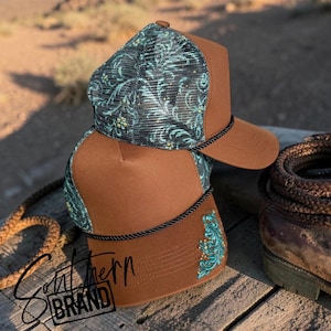 May include: A brown trucker hat featuring a teal and black floral pattern on the mesh back panels and a matching teal floral embroidery on the side. The hat includes a black braided cord across the base of the crown and a solid brown front panel.