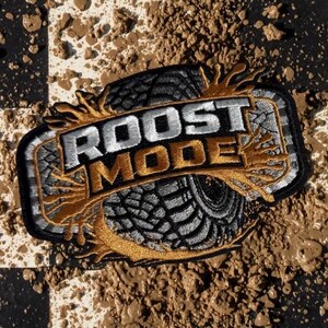 May include: An embroidered patch featuring a black tire tread design with silver and gold thread accents. The text ROOST MODE is stitched in silver and gold across the center, surrounded by stylized gold mud splatter embroidery on a black border.