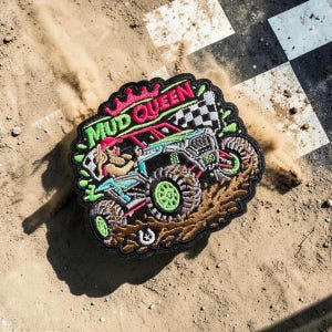 May include: Embroidered patch featuring a woman driving an off-road vehicle, with the text "MUD QUEEN" in green and pink. The patch has a black border and a checkered flag design. The vehicle is depicted in shades of blue, green, and pink.