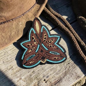 May include: Embroidered patch with a floral design in brown, black, and turquoise. The patch is on a wooden surface, with a brown cowboy hat and rope in the background. The patch has a detailed, textured appearance.