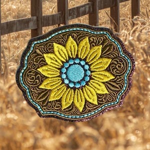 May include: An oval-shaped belt buckle featuring an embroidered sunflower design with bright yellow petals and a light blue center. The background is dark brown with intricate scrollwork patterns, bordered by a thin line of light blue stitching.
