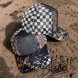 May include: Two black and white trucker hats featuring a checkered pattern on the mesh panels and a black foam front panel. Both hats are decorated with brown tire track prints and splatter designs. The text Southern BRAND is visible in black.