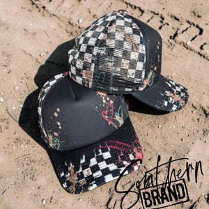 May include: Two black trucker hats featuring a white and black checkered pattern on the front panels and mesh back. The hats are decorated with paint splatters in shades of pink, teal, and tan, along with hand-drawn heart outlines and tire tracks.
