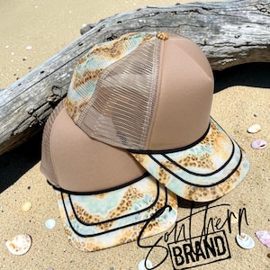 May include: Two trucker hats featuring a solid tan front panel and a tan mesh back. The brim and side panels display a light blue and tan marbled pattern with a leopard print overlay. Each brim is accented with two parallel black stitched lines.