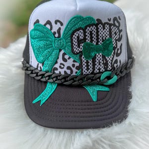 May include: A black and white trucker hat with a green glitter bow and the words "Come Day" in green glitter. The hat has a black chain around the brim.