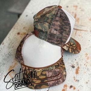 May include: Two trucker hats featuring a solid white front panel and a camouflage patterned mesh back. The brim is also camouflage with a brown trim detail. The text Southern BRAND is printed in black script and block letters on the lower left side.