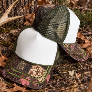 May include: Two trucker hats featuring a white front panel, olive green mesh back, and a brim with a patchwork pattern of brown, black, and pink floral and leopard prints. The hats have a green trim along the edge of the brim and front panel.