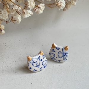May include: A pair of white ceramic cat-shaped stud earrings. Each earring has a blue floral pattern and gold-coloured accents on the ears. The earrings are displayed on a light-coloured surface, with dried white flowers in the background.