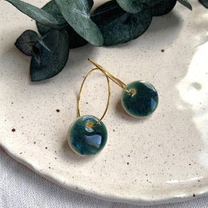 May include: Gold hoop earrings featuring dark teal ceramic beads. The beads are round and have a glossy finish. The earrings are presented on a white plate with a textured surface, with green leaves in the background.
