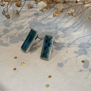 May include: A pair of rectangular ceramic stud earrings. The earrings are glazed in teal and blue, with a white border. The earrings are displayed on a white surface with gold star-shaped confetti and dried flowers.
