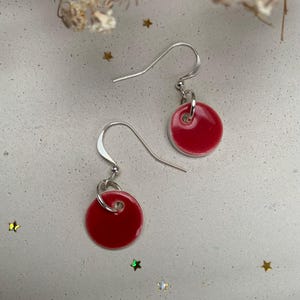 May include: A pair of red disc earrings with silver hooks. The earrings have a glossy finish and a small white border. They are displayed on a light grey surface with star-shaped decorations. The earrings are a simple, elegant design.