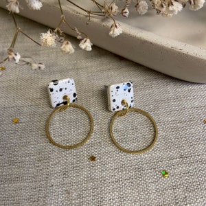 May include: A pair of earrings featuring square white tops with black speckles, connected to gold-coloured circular hoops. The earrings are displayed on a neutral surface, with star-shaped decorations and small white flowers.
