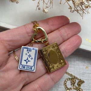 May include: A gold-colored necklace featuring a heart-shaped clasp, a small tarot card pendant with the text "THE STAR", and a gold locket. The tarot card is white with blue accents. The locket is rectangular with an ornate design.