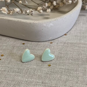 May include: A pair of light green heart-shaped stud earrings. The earrings are displayed on a textured, neutral-toned surface. A light grey, curved tray and dried flowers are in the background.
