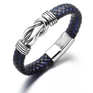 May include: A black and blue braided leather bracelet with a silver-colored knot design in the center. The bracelet has a magnetic clasp and is a stylish accessory. The braided leather is accented with blue stitching.