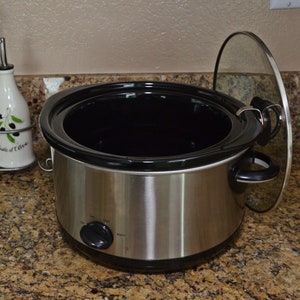 May include: A stainless steel slow cooker with a black inner pot and a glass lid. The lid is slightly ajar, revealing the empty pot.