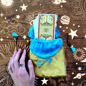 Puede incluir: Una bolsa de cordón de color dorado y azul con una carta de tarot en su interior. La bolsa está decorada con un diseño celestial y la carta muestra un árbol con un sol y una luna encima.