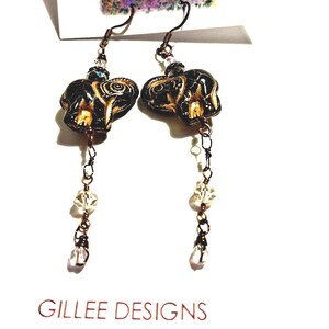 May include: A pair of dangle earrings featuring black elephant-shaped beads with gold accents. The earrings have copper-colored chains and clear crystal beads. The text "Gillee Designs" is visible at the bottom.