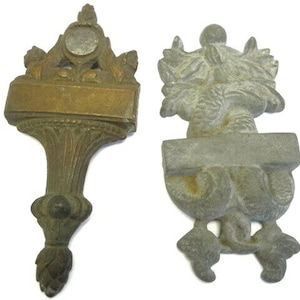 May include: Two antique metal decorative architectural elements. One is bronze-toned, the other is gray. Both have ornate designs, including floral and scrollwork details, with a blank rectangular space for text. The bronze element has a stylized finial.