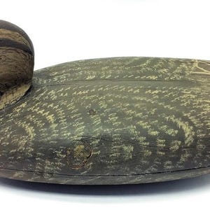 May include: A wooden duck decoy, painted in shades of brown and green. The duck has a detailed head with a dark beak and a realistic eye. The body is textured with a feather pattern, and the wings have lighter markings.