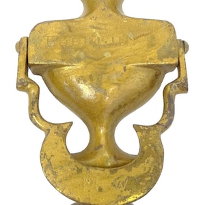 May include: A vintage brass door knocker with a weathered gold finish. The knocker features a decorative design with a floral top, a rectangular plate with text, and a curved bottom element. The overall shape is symmetrical and ornate.