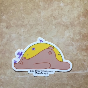 May include: A sticker featuring a cartoon bear lying down with a yellow sun background. The bear is light brown with a flower on its back. The text "The Bear Minimum (I woke up)" is below the bear.