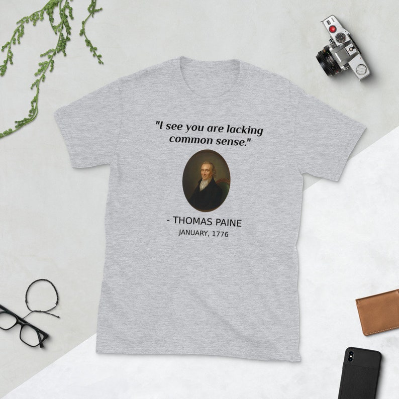 May include: A light gray t-shirt featuring a portrait of Thomas Paine and the quote "I see you are lacking common sense." The text below the portrait reads "- THOMAS PAINE JANUARY, 1776."