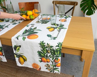 Puede incluir: Un camino de mesa de lino blanco con estampado de cítricos. El camino de mesa presenta limones, limas y naranjas en una rama verde frondosa. El camino de mesa está colocado sobre una mesa de madera con dos cubiertos y un bol de cítricos.