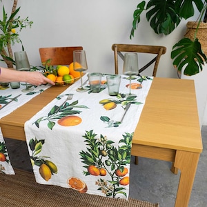 Op de afbeelding: Een witte linnen tafelrunner met een citrusfruitprint. De runner heeft citroenen, limoenen en sinaasappels op een groene, bladerrijke tak. De runner ligt op een houten tafel met twee couverts en een kom met citrusfruit.