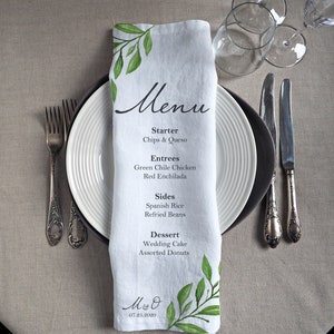 Könnte beinhalten: Eine weiße Leinen-Serviette mit einem grünen Blattdesign und dem Wort "Menu" in Schwarz gedruckt. Die Serviette listet ein Hochzeitsmenü mit Vorspeisen, Hauptgerichten, Beilagen und Desserts auf. Die Serviette enthält auch die Initialen "M & O" und das Datum "07.25.2020" in Schwarz gedruckt.