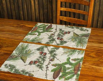 May include: Two rectangular placemats with a nature-inspired design featuring green pine branches, brown pine cones, and red berries on a cream background. The placemats are placed on a wooden table, with a wooden chair in the background.