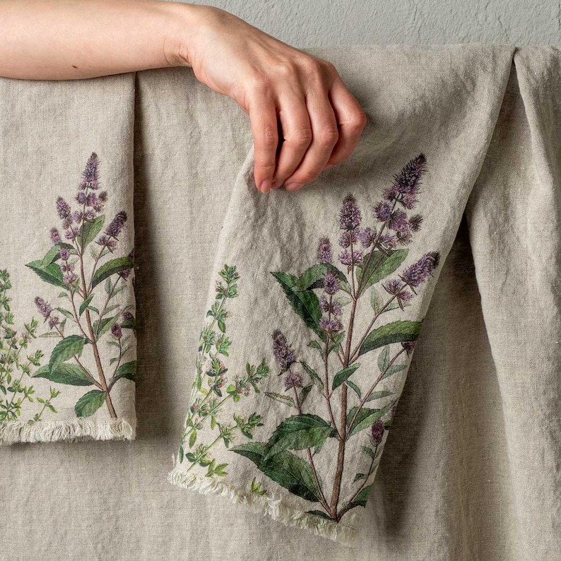 May include: Two linen tea towels with a floral print of green leaves and purple flowers. The towels have a frayed edge.