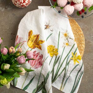 Puede incluir: Paño de cocina de lino blanco con un diseño floral de tulipanes amarillos, narcisos y tallos verdes. El paño está doblado sobre un salvamanteles tejido, con un ramo de tulipanes rosas y un cuenco de golosinas cerca.