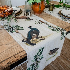 Puede incluir: Un camino de mesa de lino con fondo beige y un patrón repetido de hojas verdes, bellotas y pájaros negros y marrones. Los pájaros son un urogallo, un faisán y una agachadiza.