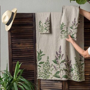 May include: Set of three linen kitchen towels with a floral print of green leaves and purple flowers. The towels are hanging on a wooden slatted wall.