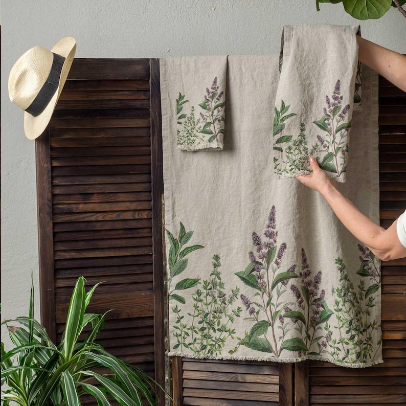 May include: Set of three linen kitchen towels with a floral print of green leaves and purple flowers. The towels are hanging on a wooden slatted wall.