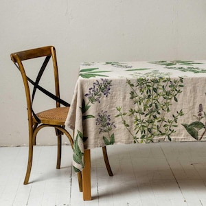 Peut inclure: Une nappe en lin beige avec un motif floral composé de feuilles vertes et de fleurs violettes. La nappe est drapée sur une table en bois avec une chaise en bois à côté.