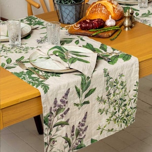 Peut inclure: Une nappe en lin beige avec des illustrations botaniques vertes d'herbes et de fleurs. Une assiette avec une serviette et des couverts sont posées sur la nappe. Une table en bois est garnie de pain, d'ail et d'épices.