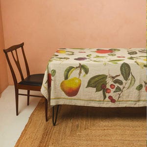 May include: A rectangular tablecloth featuring a botanical design of fruits and vegetables, including pears and cherries. The table has black hairpin legs. A wooden chair with a black seat is next to the table. The tablecloth is on a table.