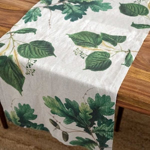 May include: A white linen table runner with a green leaf pattern. The leaves are a variety of shapes and sizes, and they are arranged in a random pattern. The runner is perfect for adding a touch of nature to your dining table.